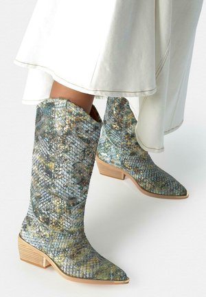 Cowboylaarzen met een patroon van blauw, groen en goud slangenleer en houten lage blokhakken gedragen bij witte wijde broekspijpen.