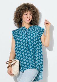 Türkise Bluse mit kurzem, rüschendem Ärmel, ausgestattet mit einem geometrischen weißen Design, kombiniert mit einer gewebten cremefarbenen Tasche und hellen Jeans.