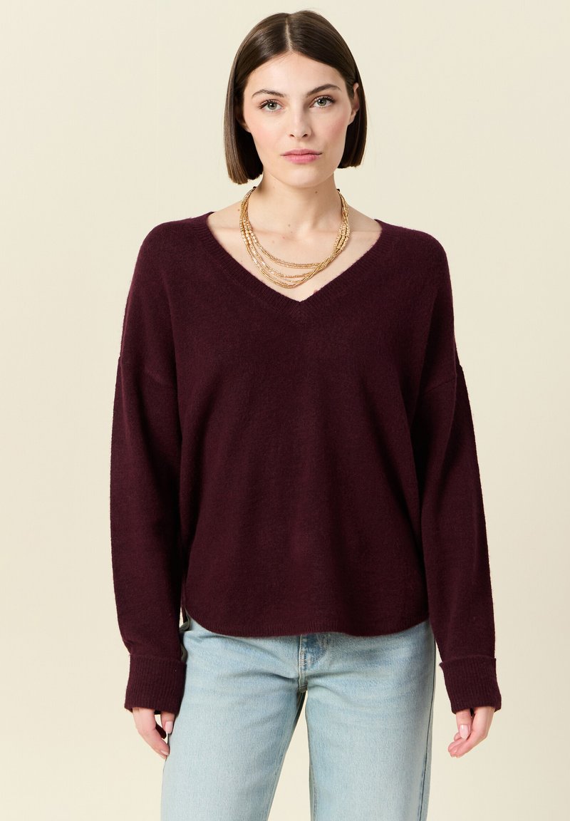 Burgundy stickad tröja med V-ringning, sänkta axlar och ribbade ärmslut; har en avslappnad passform och mjuk textur. Kombinerad med ljusblå jeans.