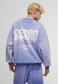 Lichtpaarse sweatshirt met een oversized pasvorm, voorzien van witte tekst op de rug, geribbelde mouwen en een zachte textuur. Gecombineerd met bijpassende joggingbroek.
