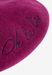 Purpura filca berete ar melnu izšuvumu kursīvā, kas uzrakstīts "Oh là là" uz baltas fona.