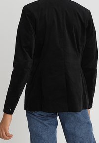 Osoba ubrana w dopasowaną czarną marynarkę i niebieskie dżinsy, widoczna z tyłu na neutralnym tle.