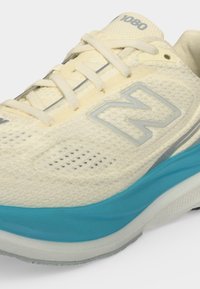Αθλητικό παπούτσι New Balance σε κρεμ χρώμα με μπλε ενδιάμεση σόλα, επάνω μέρος από δίχτυ, κορδόνια και "1080" τυπωμένο στη γλώσσα.