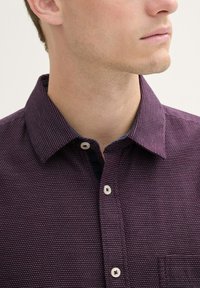 Burgundy button-up shirt with a textured pattern, contrasting collar, and white buttons. Features a single chest pocket.