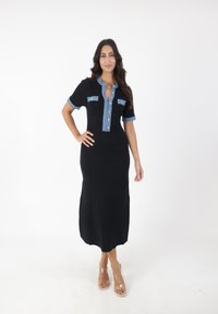 Zwarte geribbelde midi-jurk met korte denimmouwen en kraag, met een knopenlijst en twee borstzakken. Heldere sandalen.