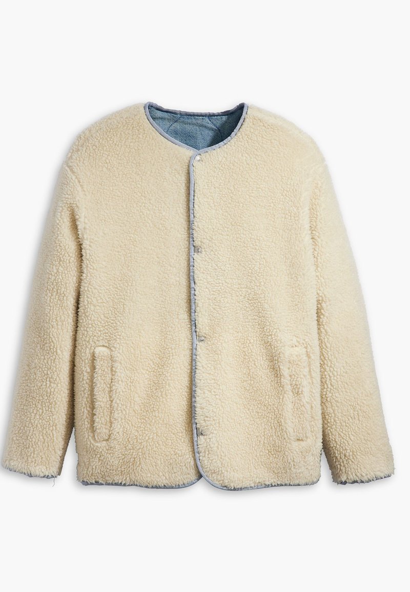 Chaqueta de forro polar color crema con cuello redondo, botones a presión y dos bolsillos laterales. Ribeteada con tela vaquera en los bordes.