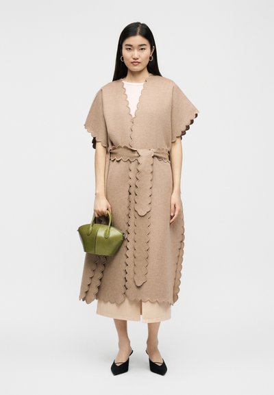 Γυναίκα που φοράει ένα μπεζ παλτό με ζώνη και κυματιστό τελείωμα, κρατώντας μια μικρή τσάντα ελιάς, συνδυασμένη με μαύρες γόβες και ανοιχτόχρωμο παντελόνι.