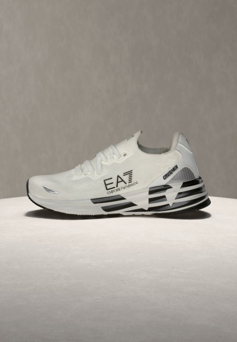 Бял маратонка Emporio Armani EA7 с черна райета подметка и текст "Crusher" на петата, поставена на светла повърхност на сив фон.