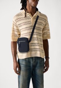 Polo shirt all'uncinetto beige con maniche corte e vestibilità ampia, abbinato a una borsa a tracolla blu scuro con zip e logo. Jeans in denim scuro.