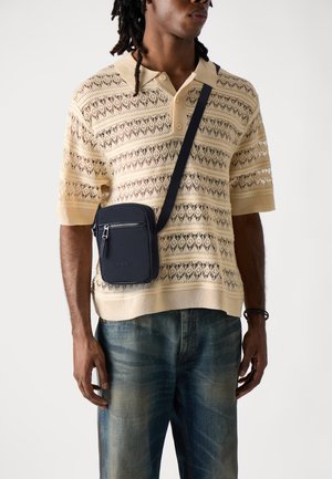 Beige gehäkeltes Poloshirt mit kurzen Ärmeln und lockerer Passform, kombiniert mit einer dunkelblauen Schultertasche mit Reißverschluss und Logo. Dunkelblaue Jeans.