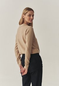 Beige Strickpullover mit umgeschlagenem Kragen, kombiniert mit dunklen, strukturierten Hosen. Das Modell ist leicht zur Seite gedreht und zeigt die Rückansicht des Outfits.