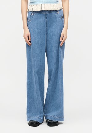 Donna che indossa jeans a gamba larga a vita alta di colore blu con dettagli a bottone sulle tasche anteriori, abbinati a scarpe nere e a una canottiera a righe senza maniche.