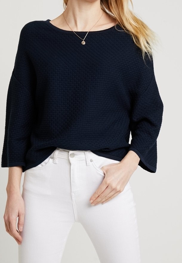 Femme portant un haut texturé bleu marine à manches longues avec un large décolleté et un jean blanc taille haute, la main reposant près de la poche avant.