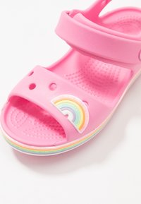 Vaaleanpunaiset muoviset sandaalit, joissa on avoin design, säädettävät hihnat ja sateenkaaren aksentti. Texturoitu pohja ja värikäs reunan viimeistely.