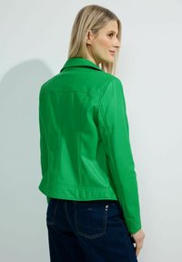 Veste en simili cuir vert au design ajusté, avec un col à pression et une silhouette cintrée, associée à un pantalon en denim bleu foncé.