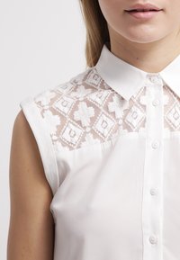 Blusa bianca con colletto, maniche corte e schiena in pizzo con motivi floreali. Chiusura a bottoni lungo il fronte. Tessuto dalla texture liscia.