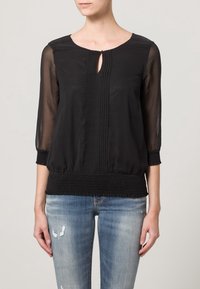 Svart genomskinlig blus med trekvartsärmar, nyckelhålsklippning, vertikala veck och samlad elastisk fåll. Kombinerad med slitna ljusa jeans.