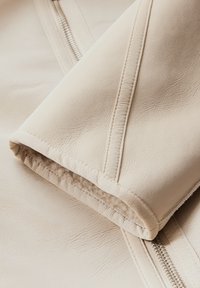 Beige imitationläderjacka med synliga sömmar, texturerad yta och dragkedja. Kragen och ärmsluten har ett mjukt innerfoder.