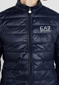 Námořnická modrá péřová bunda s vysokým límcem, zapínáním na zip, horizontálním prošíváním a logem EA7 Emporio Armani na levé části hrudi. Hladká textura.