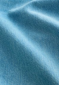 Tissu en denim bleu clair avec texture tissée diagonale visible et plis doux créant des ombres délicates.