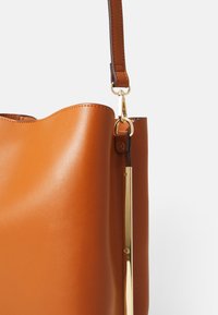 Brun læderhåndtaske med en glat tekstur, guldfarvet hardware og et karakteristisk bølget kantdesign øverst. Inkluderer en aftagelig rem.