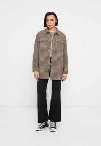 Καφέ και λευκό καρό υπερμεγέθες πουκάμισο-μπουφάν με κουμπιά, εμπρόσθιες τσέπες και άνετη εφαρμογή, συνδυασμένο με μαύρο παντελόνι φαρδιάς γραμμής και αθλητικά παπούτσια.