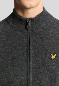 Grijze zip-up sweater met een hoge kraag, met een subtiele textuur en een klein geel geborduurd logo op de linkerkant van de borst.