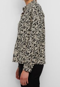 Femme portant un chemisier à manches longues noir et beige à motifs floraux avec un col à volants, associée à un pantalon noir, debout de profil.