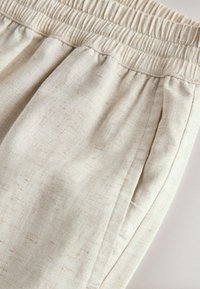 Pantalon en lin beige avec taille élastique et poche latérale, montrant la texture du tissu et les coutures cousues.