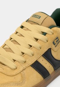 Baskets en daim jaune avec des bandes latérales noires, des lacets jaunes épais et un col matelassé vert, arborant le logo de la marque Globe sur la languette et le côté.