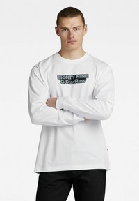 Hvit langermet skjorte med rund hals, med et svart grafisk trykk som leser "EIGHTY NINE G-StarRaw" over fronten.