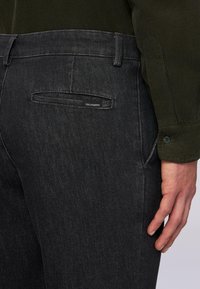 Ingrandimento della mano di una persona che riposa accanto al fianco, indossando pantaloni grigio scuro con un'etichetta Trussardi vicino alla tasca posteriore e una maglia verde scuro.
