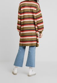Proužkovaný pletený cardigan v olivově zelené, růžové, červené a tmavě modré barvě, kombinovaný s světlými modrými rozšířenými džínami a bílými teniskami. Žebrovaná textura, volný střih.