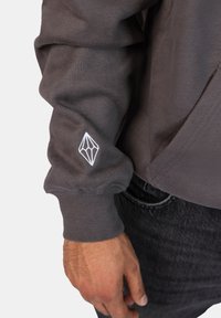 Felpa grigia con polsini a costine, con un logo geometrico bianco sulla manica. Il materiale sembra morbido con una texture liscia.