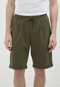 Uomo che indossa pantaloni corti casual verde oliva con vita regolabile e orli arrotolati, abbinati a una maglietta bianca senza maniche.