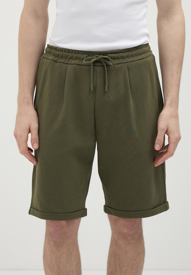 Uomo che indossa pantaloni corti casual verde oliva con vita regolabile e orli arrotolati, abbinati a una maglietta bianca senza maniche.