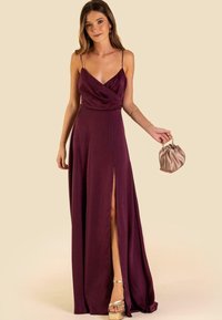 Lungo abito prugna con davanti drappeggiato, spalline sottili e spacco alta coscia. Accompagnato da una clutch in satin beige e tacchi a piattaforma metallici.