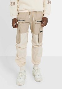 Beige cargo-byxor med kontrasterande ljusbeige paneler, flera dragkedjefickor, justerbar snörning i midjan och elastiska mudd.