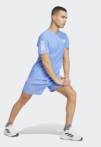 Ljust blå atletisk tröja med korta ärmar, med tre vita ränder på vänster axel, ihop med matchande shorts och vita sneakers.