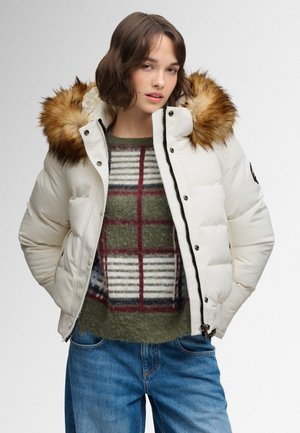 Woman wearing a white puffer jacket with fur hood, green plaid sweater, and blue jeans, standing with hands in pockets against plain background.