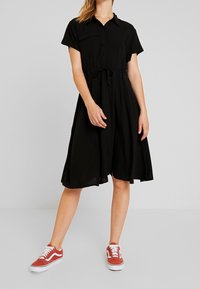 Czarna sukienka z krótkimi rękawami, z guziczkami, ze sznurkiem w talii i zwiewnym dołem, zestawiona z czerwonymi sneakersami. Gładki materiał, casualowy design.