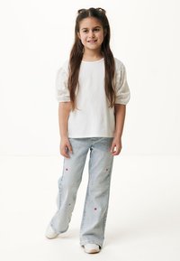 Camiseta blanca de algodón con mangas abullonadas de ojales, combinada con jeans de color azul claro con acentos bordados en rojo. La modelo sonríe de pie.