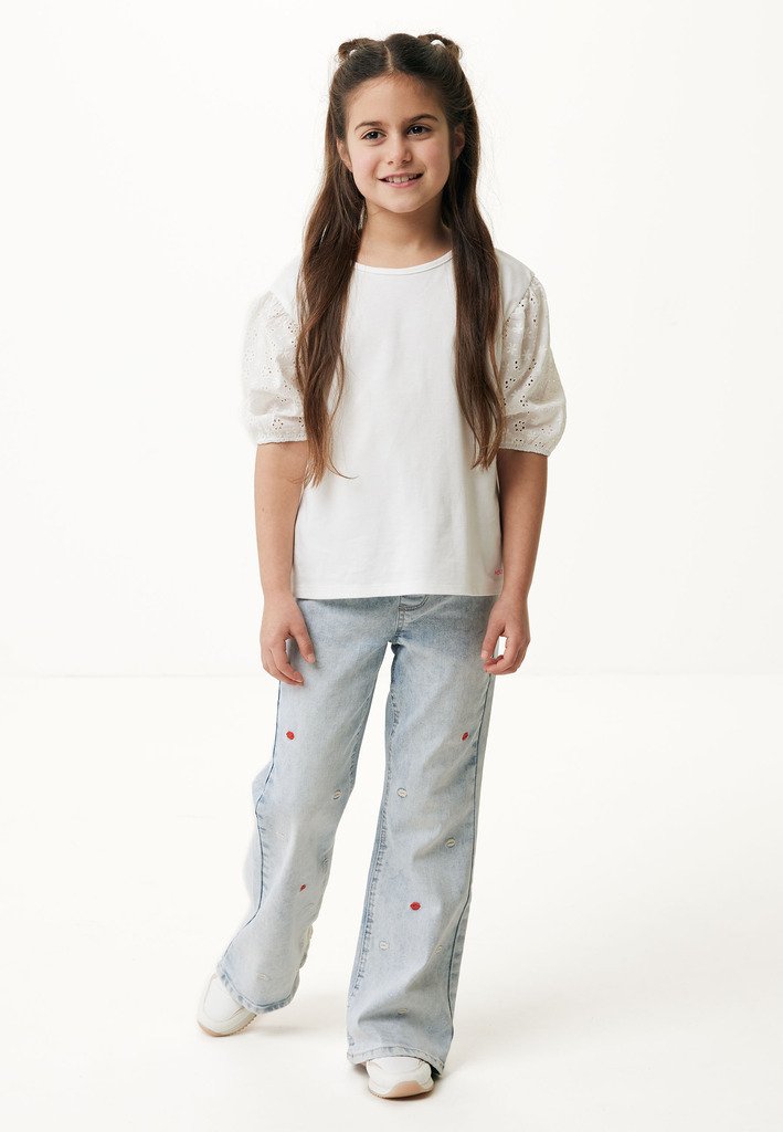 Camiseta blanca de algodón con mangas abullonadas de ojales, combinada con jeans de color azul claro con acentos bordados en rojo. La modelo sonríe de pie.