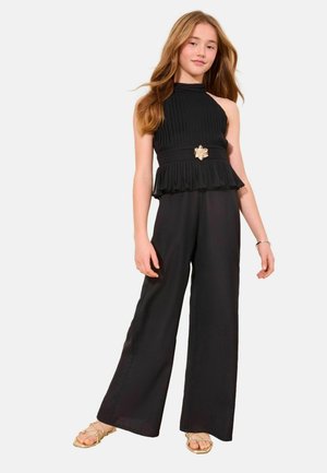 Niña joven con cabello largo, vestida con un top negro sin mangas plisado con un cinturón y pantalones negros de pierna ancha, de pie con sandalias doradas.