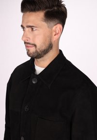 Veste en daim noir avec un col pointu, deux boutons et une poche poitrine. Texture lisse avec des variations tonales subtiles.