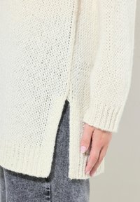 Pull en tricot surdimensionné crème avec un ourlet et des poignets côtelés. Fentes latérales pour plus de détail. Porté sur un pantalon gris, mettant en valeur la texture et l'ouverture.