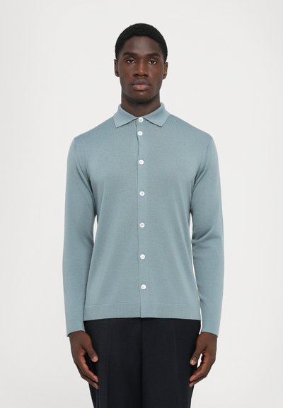 Homme portant un cardigan bleu clair à manches longues et des pantalons noirs, se tenant devant un fond blanc uni.