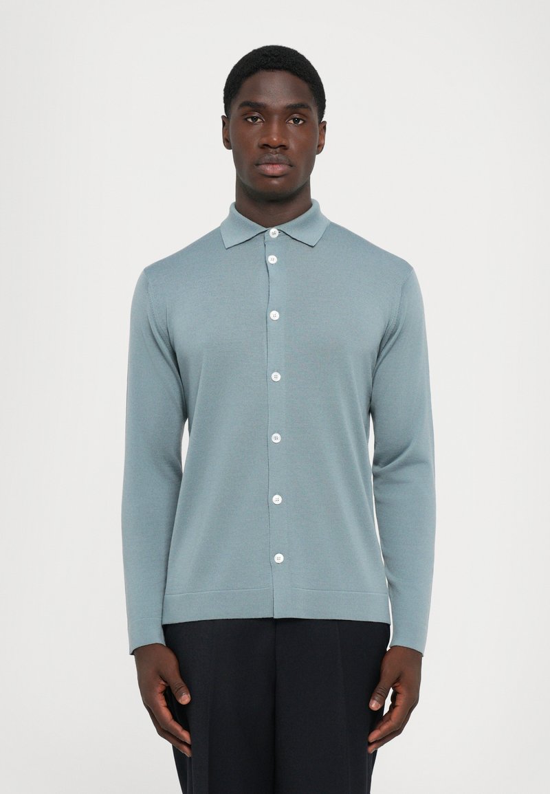 Homem com um cardigan azul claro de mangas compridas e botões, e calças pretas, de pé contra um fundo branco liso.