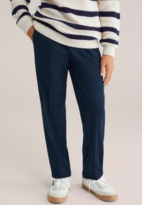 Tamno plave hlače s opuštenim krojem, izrađene od mekog materijala. Uparene s bijelim tenisicama s sivim detaljima i svijetlosmešanim potplatom.