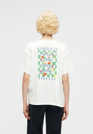 Blond krullend persoon van achteren gezien, draagt een wit T-shirt met een kleurrijke afbeelding van sardines, citroen en tomaat, met de tekst "Sardinas Frescas".